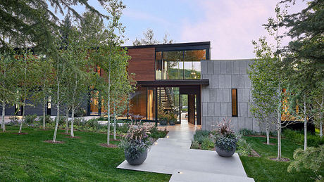 A modern, two-story home with glass walls, wood siding, and a concrete exterior, surrounded by lush greenery.