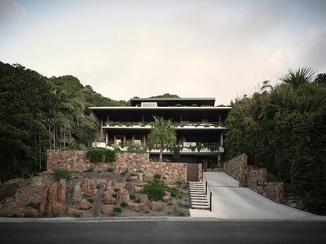 A modern, multi-story building nestled among lush greenery, featuring stone walls and balconies.