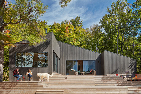 A modern, black-clad cabin nestled in a lush, autumnal forest, with a large deck and ample windows.