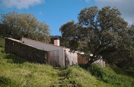 Rustic, stone-walled rural building with wooden doors, surrounded by lush vegetation.