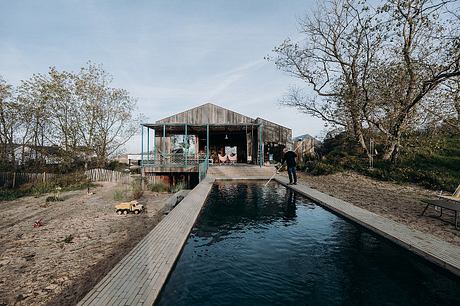 A rustic wooden cabin with a covered porch sits alongside a wooden dock over a tranquil pool.