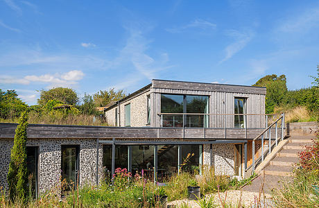 A modern, wooden house with large windows surrounded by lush greenery and a natural landscape.