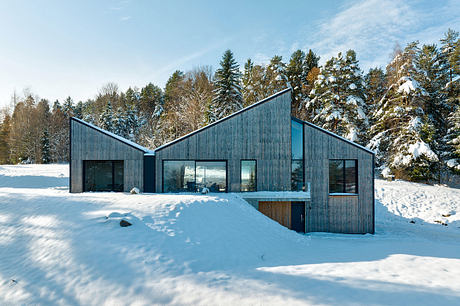 A modern, multi-gabled wooden house nestled in a snowy winter landscape, with large glass windows.