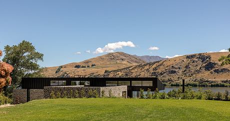 Modern lakeside home with stone walls, large windows, and grassy lawn against mountain backdrop.