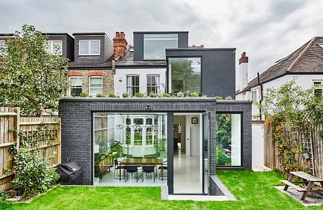 A modern, minimalist home extension with large glass windows, brick exterior, and lush greenery.