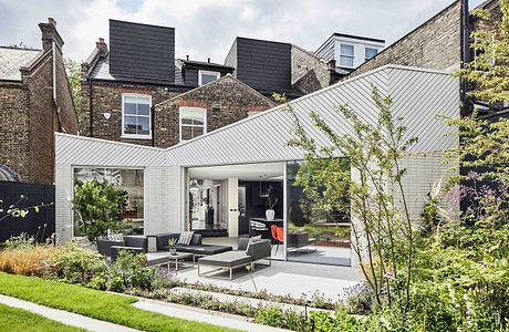 Modern glass-enclosed extension with slate roof and brick facade, surrounded by lush greenery.