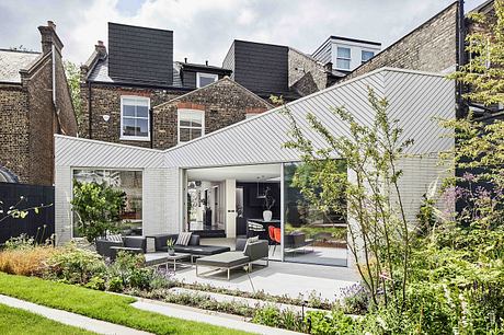 Modern glass-enclosed extension with slate roof and brick facade, surrounded by lush greenery.