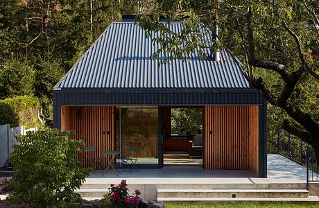 A modern, gabled cabin with a corrugated metal roof and wooden cladding, surrounded by lush greenery.