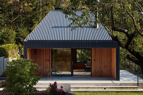 A modern, gabled cabin with a corrugated metal roof and wooden cladding, surrounded by lush greenery.