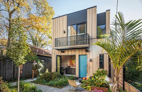 Modern two-story home with wooden siding, balcony, and lush greenery surrounding the entrance.