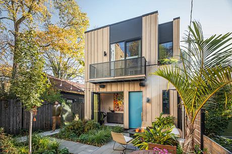 Modern two-story home with wooden siding, balcony, and lush greenery surrounding the entrance.