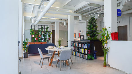 A modern, industrial-style office space with open seating areas, bookshelves, and potted plants.