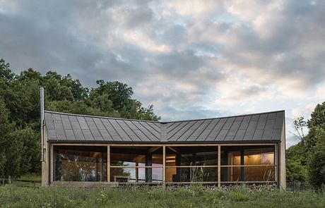 A modern cabin-like structure with a slanted metal roof and large glass windows overlooking a grassy field.