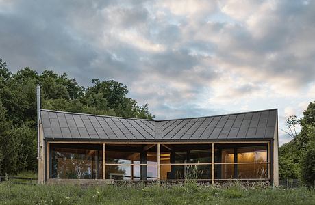 A modern cabin-like structure with a slanted metal roof and large glass windows overlooking a grassy field.