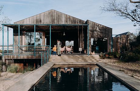 Rustic wooden cabin with open porch, pool, and reflection in water against blue sky.