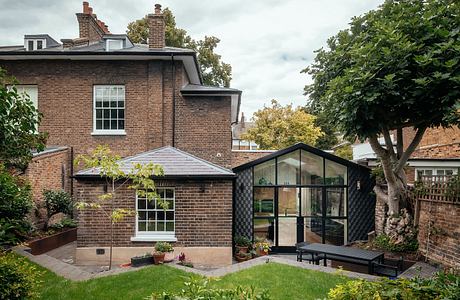 A charming brick house with a modern glass-walled extension, surrounded by lush greenery.