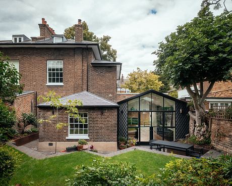 A charming brick house with a modern glass-walled extension, surrounded by lush greenery.