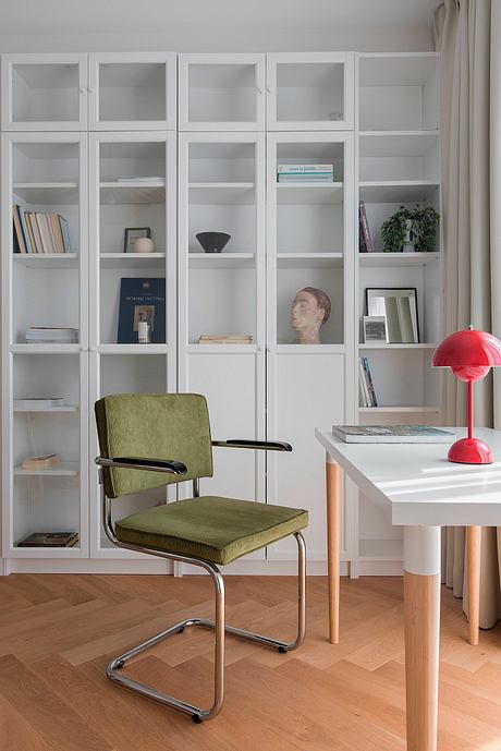 A modern, minimalist home office with built-in bookshelves, a green armchair, and a red lamp.
