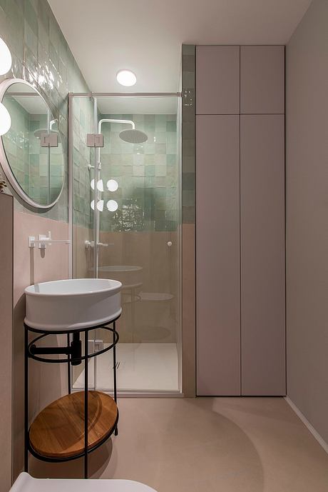 A modern bathroom with mint green tiles, a circular mirror, and built-in storage cabinets.