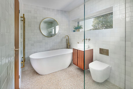 Modern bathroom with freestanding tub, round mirror, and wood vanity against wall tiles.