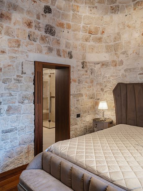 Rustic stone walls, wood-framed door, quilted bedspread, and a nightstand lamp create a cozy, intimate space.