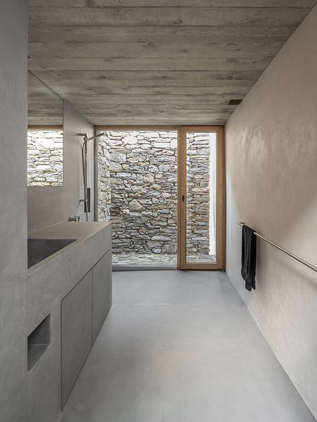A modern bathroom with concrete floors, stone walls, and a wooden door leading to the exterior.