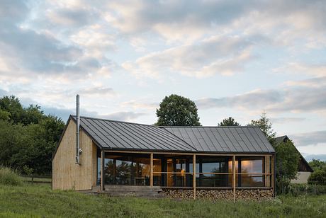 A contemporary wooden cabin with a metal roof, surrounded by lush greenery and a cloudscape.