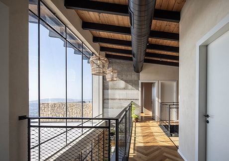 An open, airy hallway with wooden beams, a wire railing, and expansive windows overlooking a scenic view.