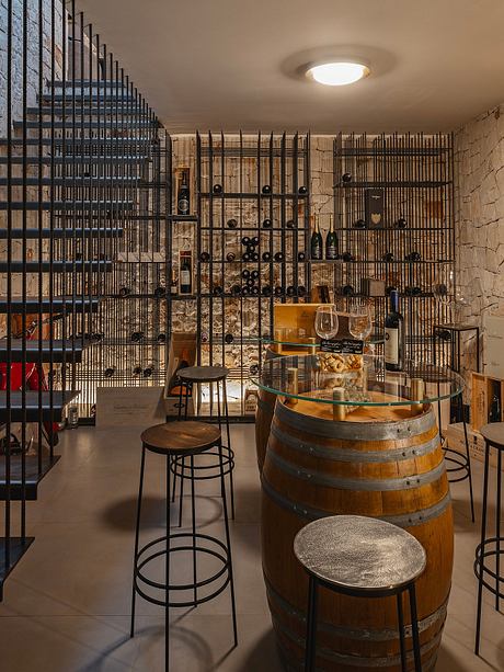 Rustic wine cellar with metal caged shelving, wooden barrels, and a glass-topped table.