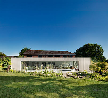 A modern glass-walled home with a traditional wooden structure above, surrounded by lush greenery.