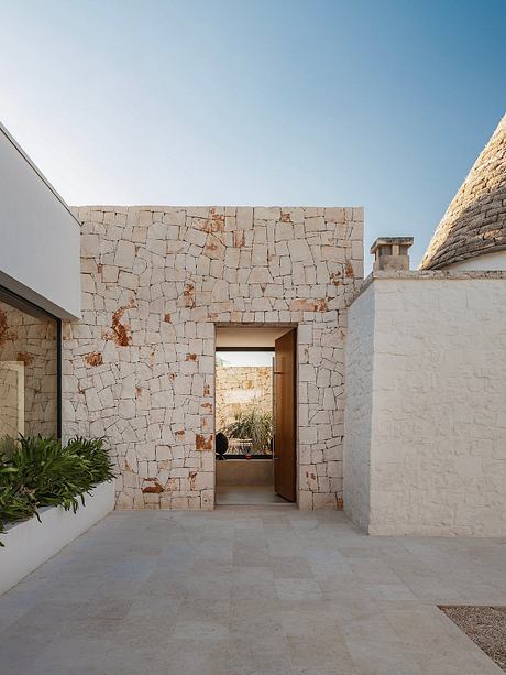 The image depicts a modern architectural design featuring a stone wall with a recessed doorway, leading to a minimalist interior space.