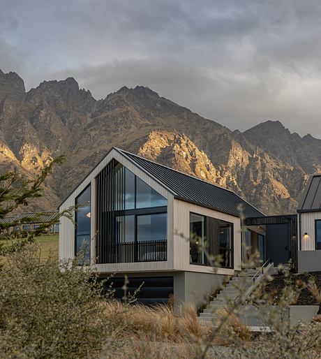 A modern cabin with a black metal roof and expansive glass walls, set against a mountainous backdrop.