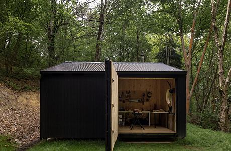 A small, black-colored shed nestled in a lush, green forest, with a glimpse of its wooden interior exposed.