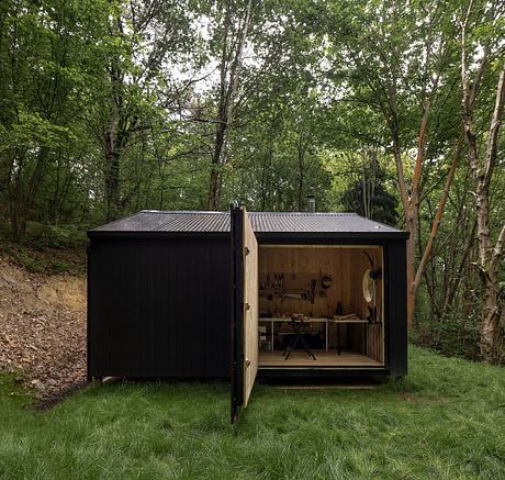 A small, black-colored shed nestled in a lush, green forest, with a glimpse of its wooden interior exposed.