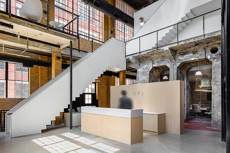 An open, industrial-style office space with exposed brick walls, metal staircase, and geometric furniture.