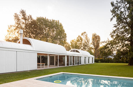 A modern, glass-enclosed home with a swimming pool and lush green surroundings.