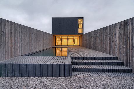 A modern, minimalist house with a dark exterior, wooden decking, and a glass-enclosed interior.