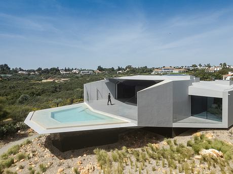 Modern, minimalist villa with geometric forms, glass walls, and a swimming pool overlooking a lush landscape.