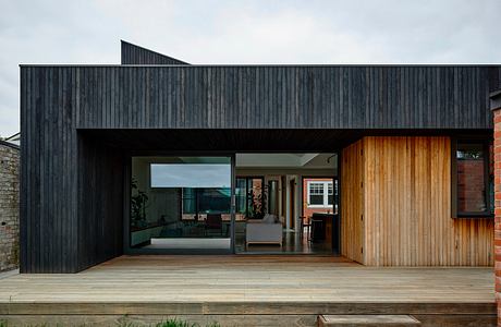 A modern architectural design featuring a dark wooden exterior, large windows, and a raised deck.