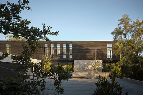 A modern, wood-paneled building with large windows and a covered entrance, surrounded by lush greenery.