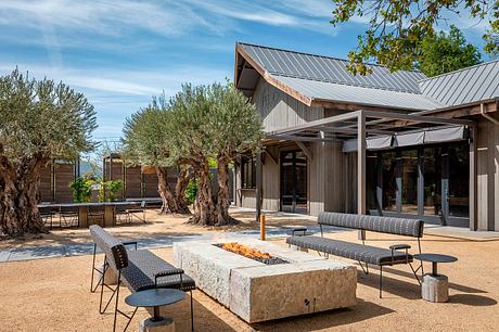 Rustic outdoor seating area with wooden building, metal roof, and olive trees.