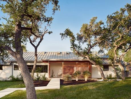 A modern, single-story house with a metal roof, surrounded by lush oak trees. The exterior features a mix of wood and concrete elements.