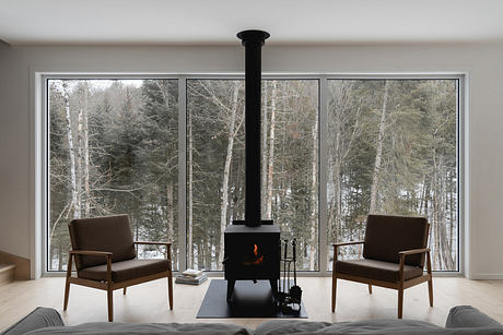 A modern, minimalist living room with large windows overlooking a forested landscape and a central fireplace.