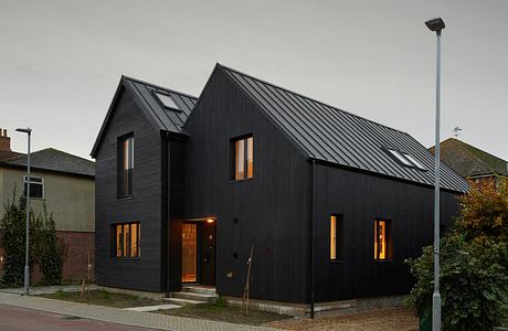 A modern, dark-colored house with triangular roof and large windows, situated on a street.