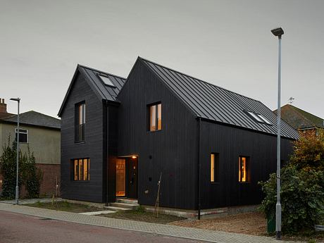 A modern, dark-colored house with triangular roof and large windows, situated on a street.