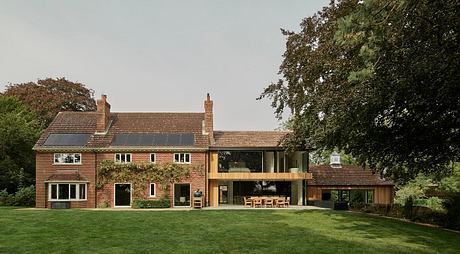 A modern two-story brick house with large windows and a glass extension, surrounded by lush greenery.