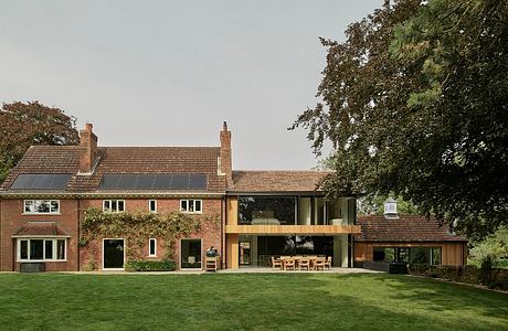 A modern two-story brick house with large windows and a glass extension, surrounded by lush greenery.