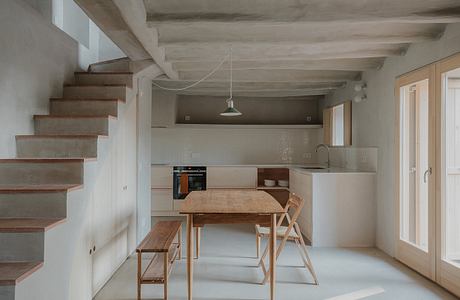 Minimalist kitchen-dining area with wooden furniture, concrete beams, and a spiral staircase.