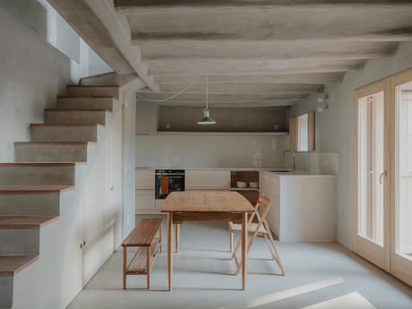 Minimalist kitchen-dining area with wooden furniture, concrete beams, and a spiral staircase.