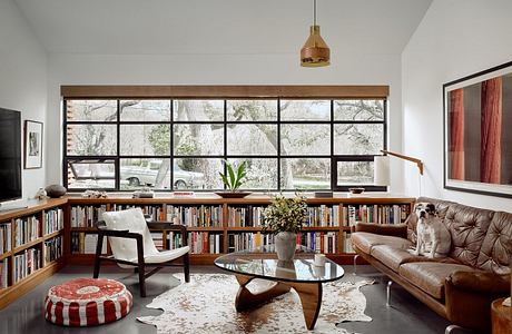 Spacious living room with wooden shelves, textured rug, and large windows showcasing outdoor greenery.